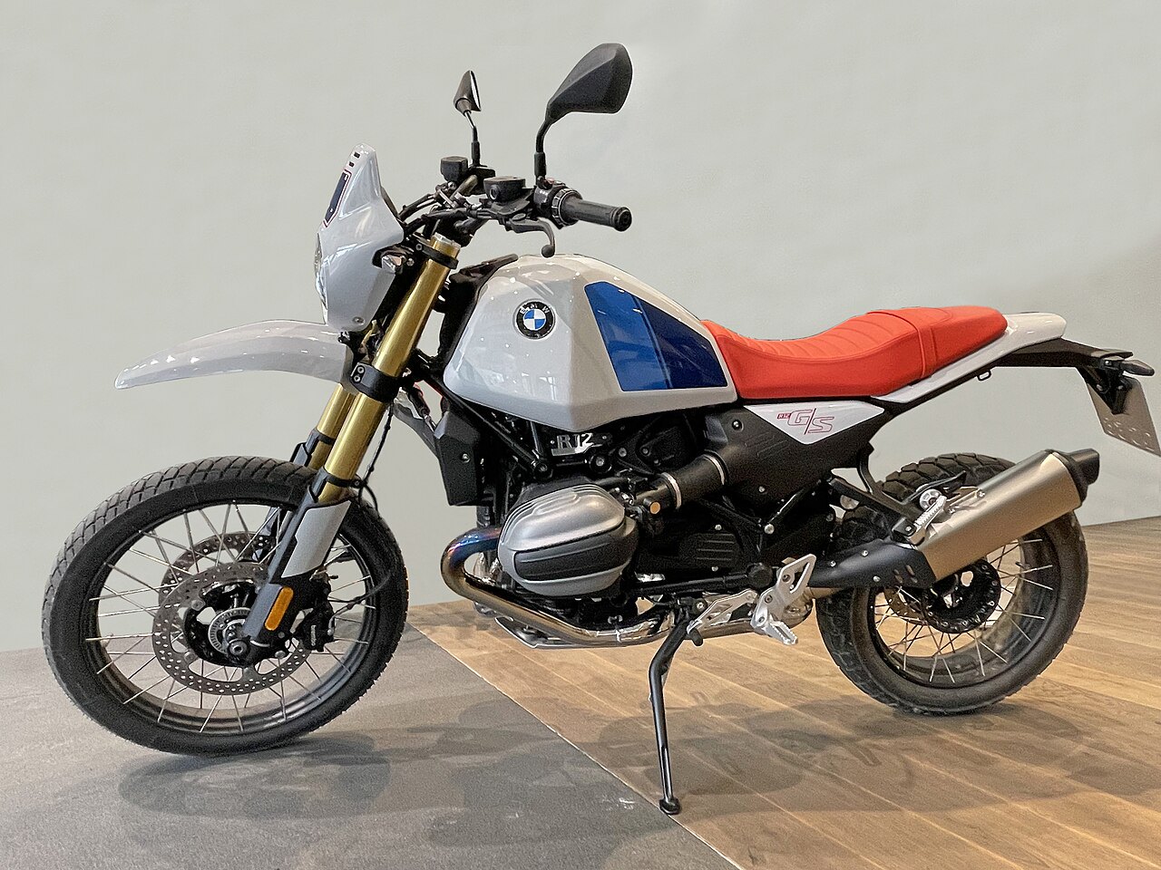 אופנוע BMW R 1250 GS בצבע אפור עם פס כחול וספסל אדום, צולם בזווית צדית על רקע לבן ורצפת עץ