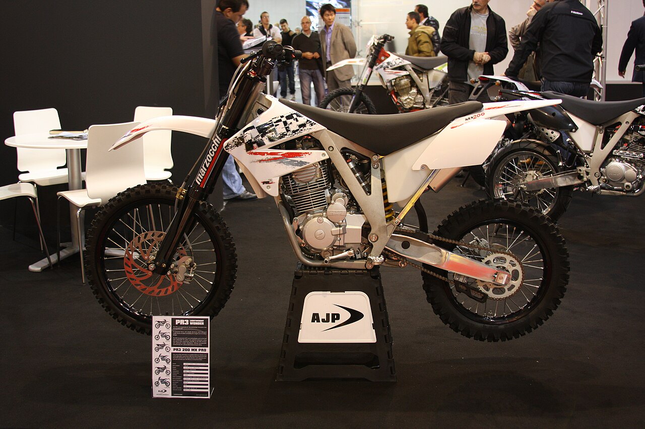 אופנוע מרוץ לבן עם מוט Marzocchi וסידור AJP, דגם PR3 200 MX PRO, בתצוגה בתערוכה עם אנשים ברקע.