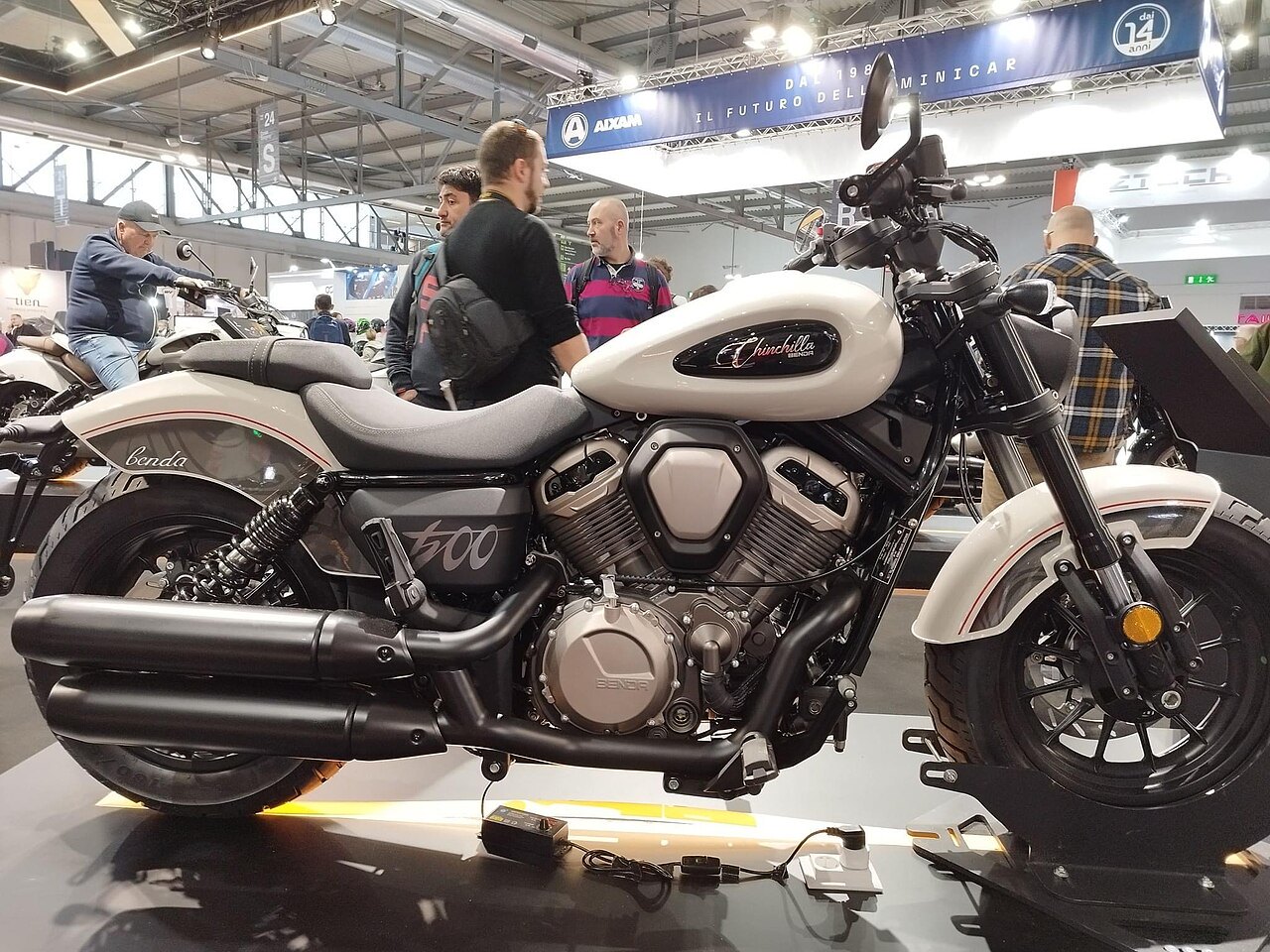 אופנוע Benelli Chinchilla 500 בצבע לבן-אפור, צולם מזווית צדית בקרוס-הול, עם אנשים בקרבתו וביתר רקע של תערוכה.