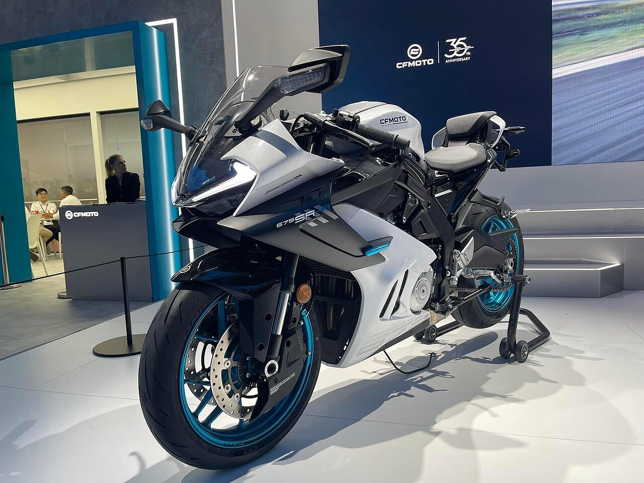 אופנוע CFMOTO 675SR-X לבן עם גלגלים כחולים, צולם בזווית צדית ביציע תצוגה עם רקע כחול וסמל 35 שנה למותג