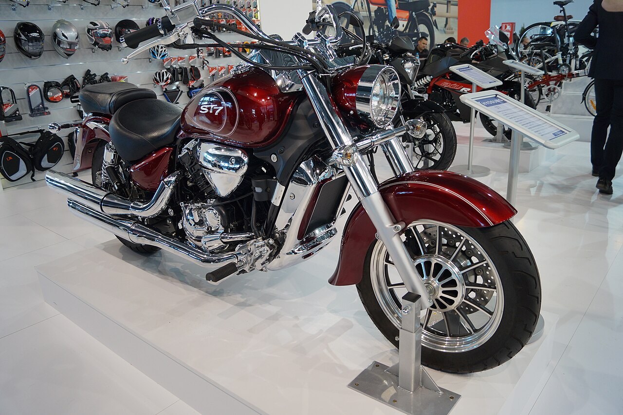 אופנוע קלאסי אדום עם מנוע כרום וסילינדר S7, צולם בזווית צדית על פלטפורמה לבנה במعرض, עם אביזרי רכיבה ברקע.