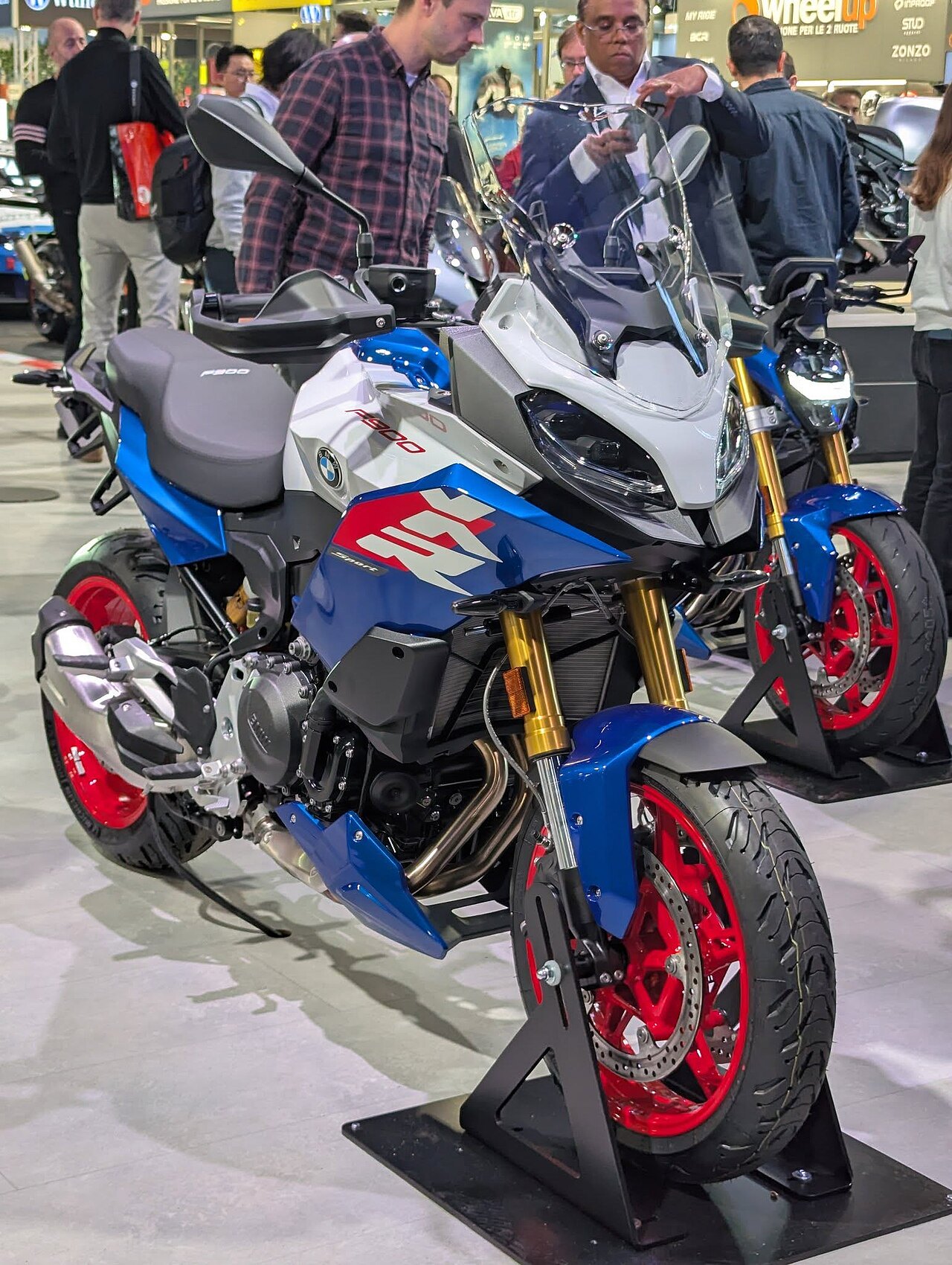 אופנוע BMW F800GS בצבע כחול, לבן ורød, מוצג בתצוגה פנימית עם גלגלים אדומים וסיבובים זהובים, סביבה של תערוכה עם מבקרים.