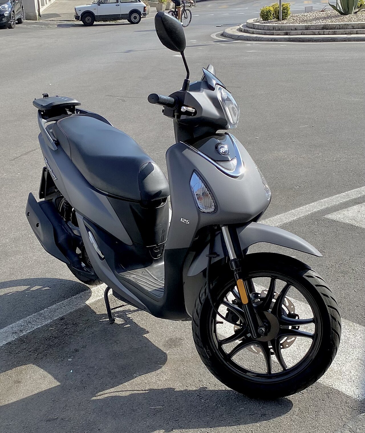 אופנוע סקוטר SYM 125 בצבע אפור כהה, צילום מזווית קדמית-צדדית, חנייה על אספלט עם רחוב ומכוניות ברקע