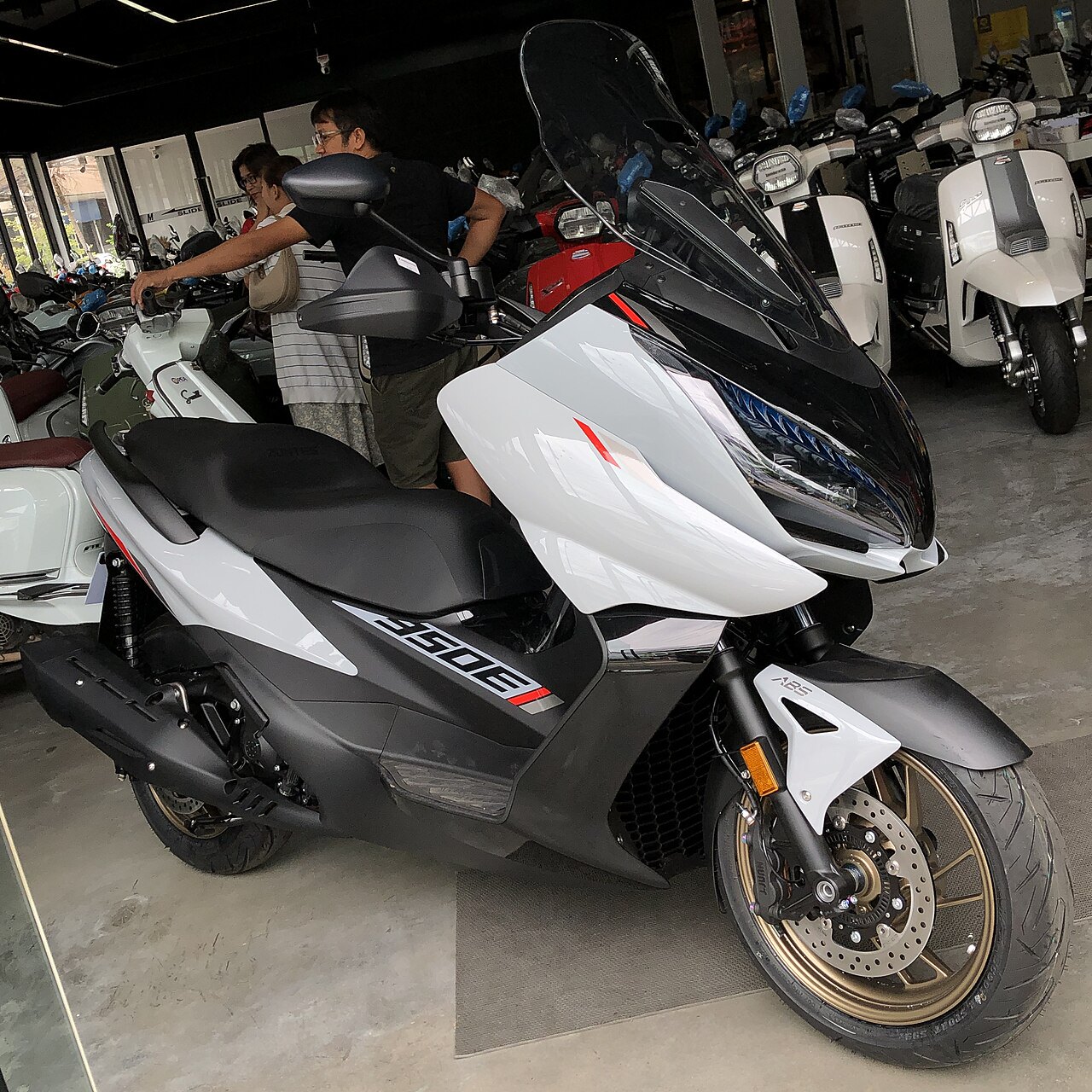 אופנוע חשמלי בצבע לבן וסגול עם סיבוב 600 על הצד, צילום קרוב מזווית צד, בתוך חנות אופנועים עם אנשים ברקע.