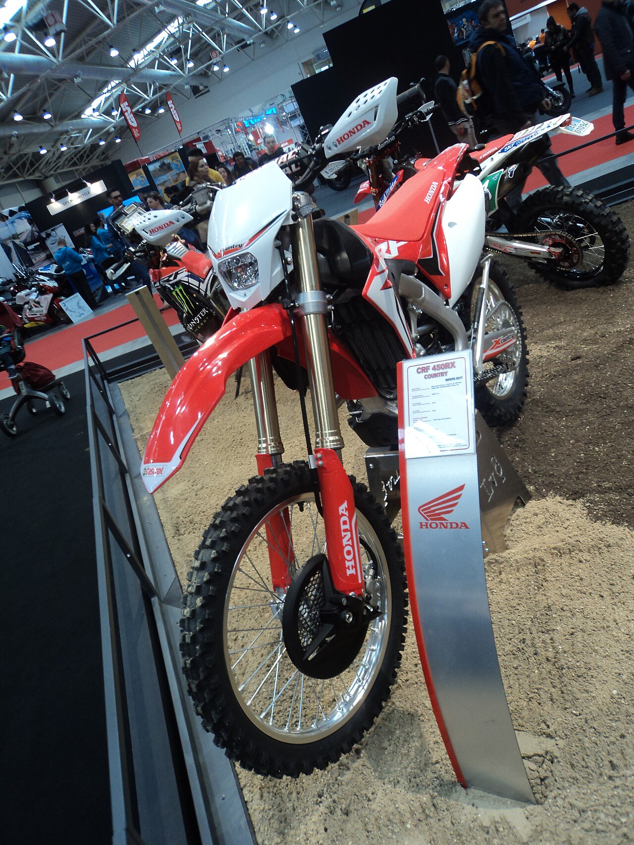 אופנוע הונדה CRF 450RX אדום-לבן בתצוגה בתערוכה, זווית קדמית עם גלגל קדמי וסימן הונדה, סביבה פנימית עם מבקרים.