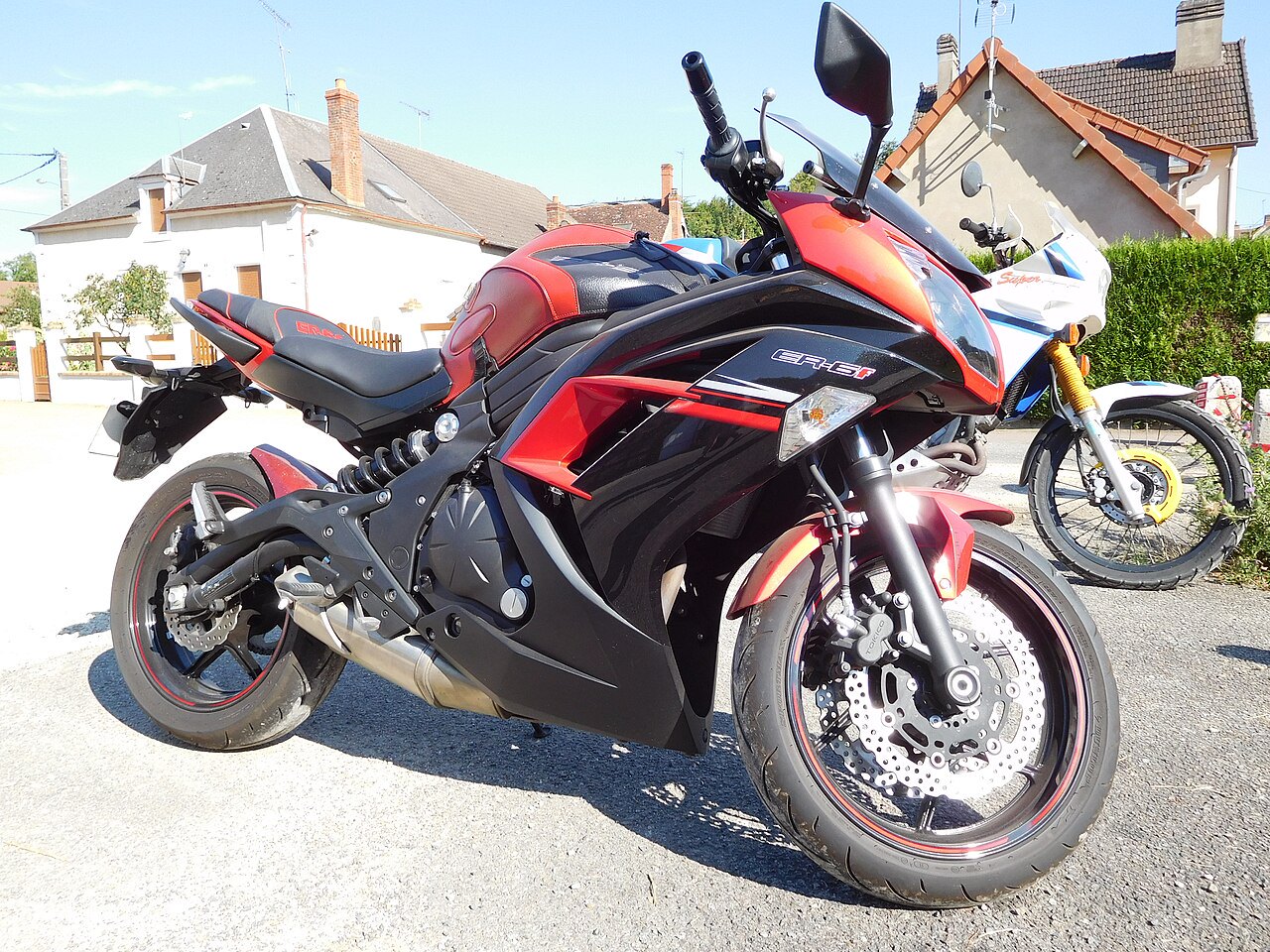 אופנוע Kawasaki ER-6f בצבעים אדום ושחור, צילום צד מקרוב על רצפת אספלט, ברקע בתים וצמחייה