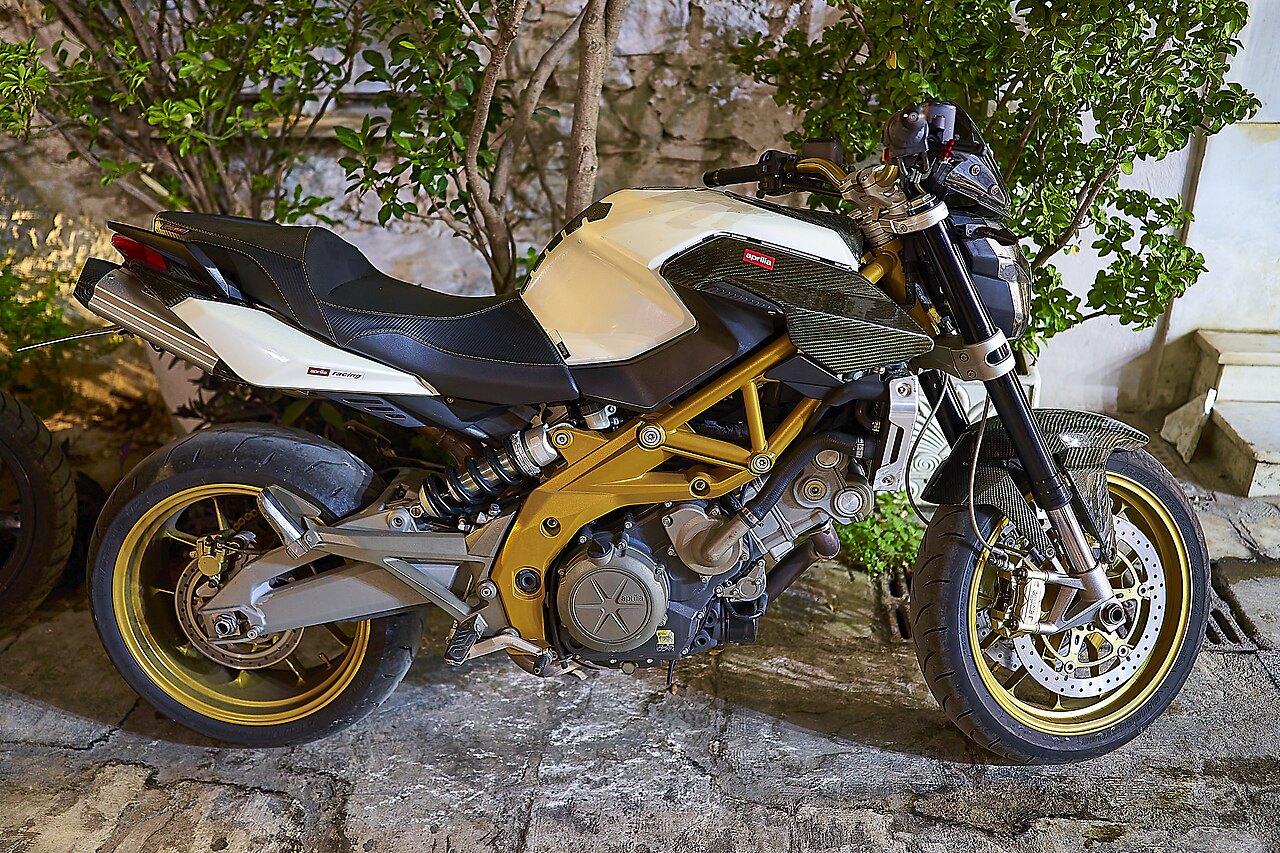 אופנוע Aprilia RSV4 Racing לבן-שחור עם מסגרת זהב, צילום צד מקרוב על רקע קיר אבן וצמחייה
