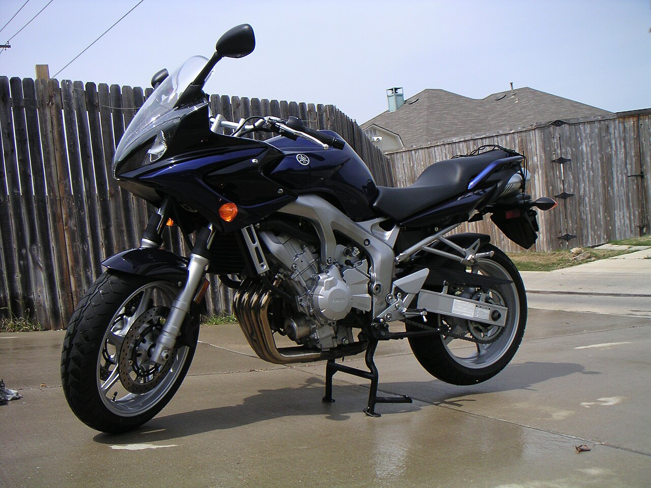 אופנוע ימבה FZ1 בצבע כחול-שחור, צילום צד, עמיד על רצפת בטון מול גדר עץ, מראה מנוע ומסגרת מטאלית