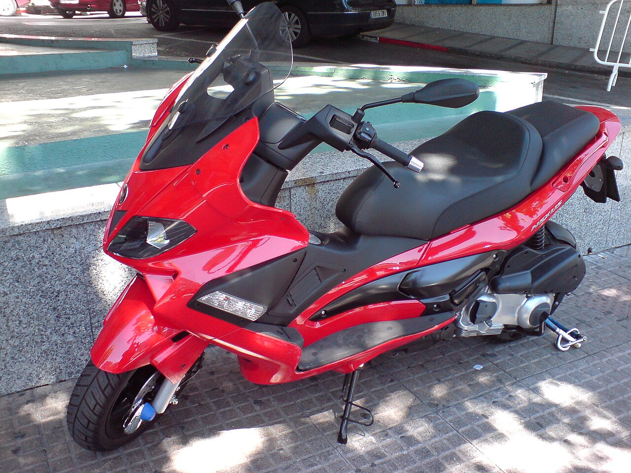 אופנוע סקוטר אדום עם מושב שחור, דגם Yamaha TMAX, צולם מזווית צד-קדימה על רצפת אבן, ברקע חומרים בניין ומכוניות