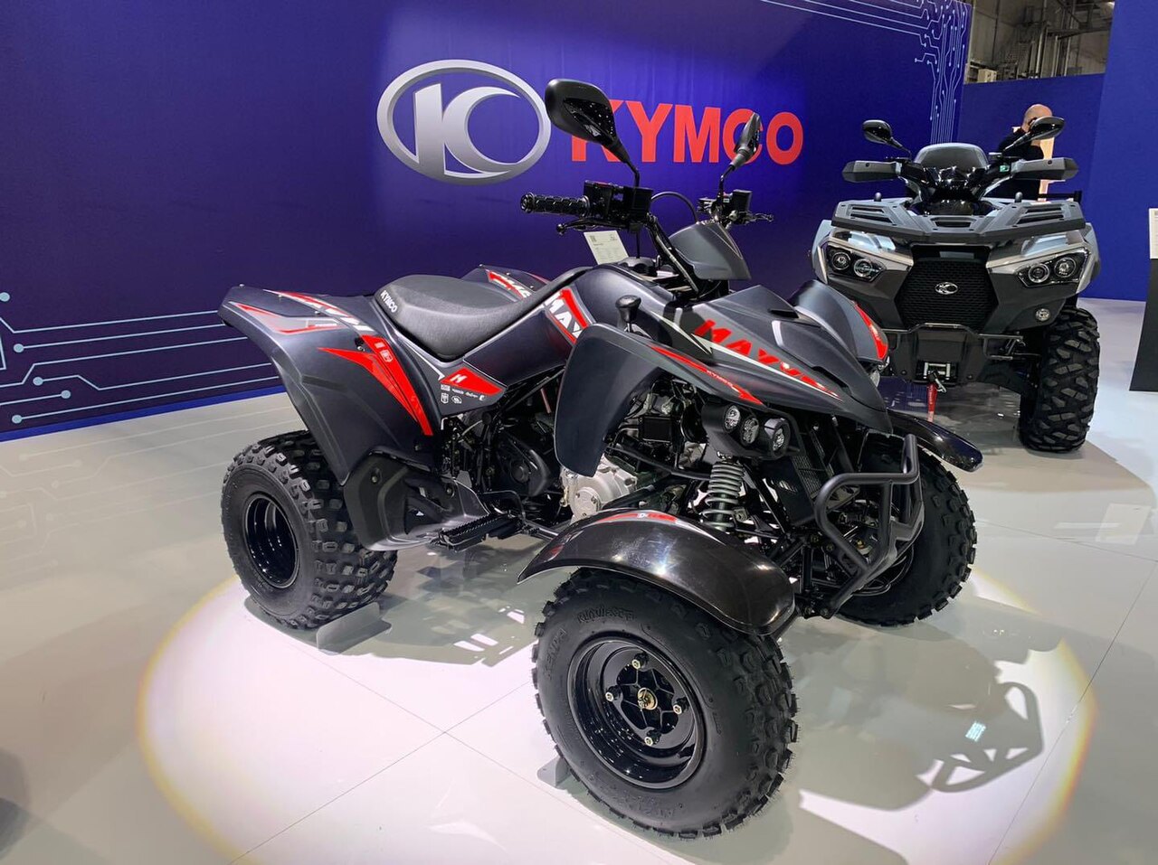 אופנועי שטח Kymco K-X4 בצבע אפור עם פסים אדומים, מוצגים בתצוגה עם רקע כחול ולוגו Kymco, זווית קדמית מימין.