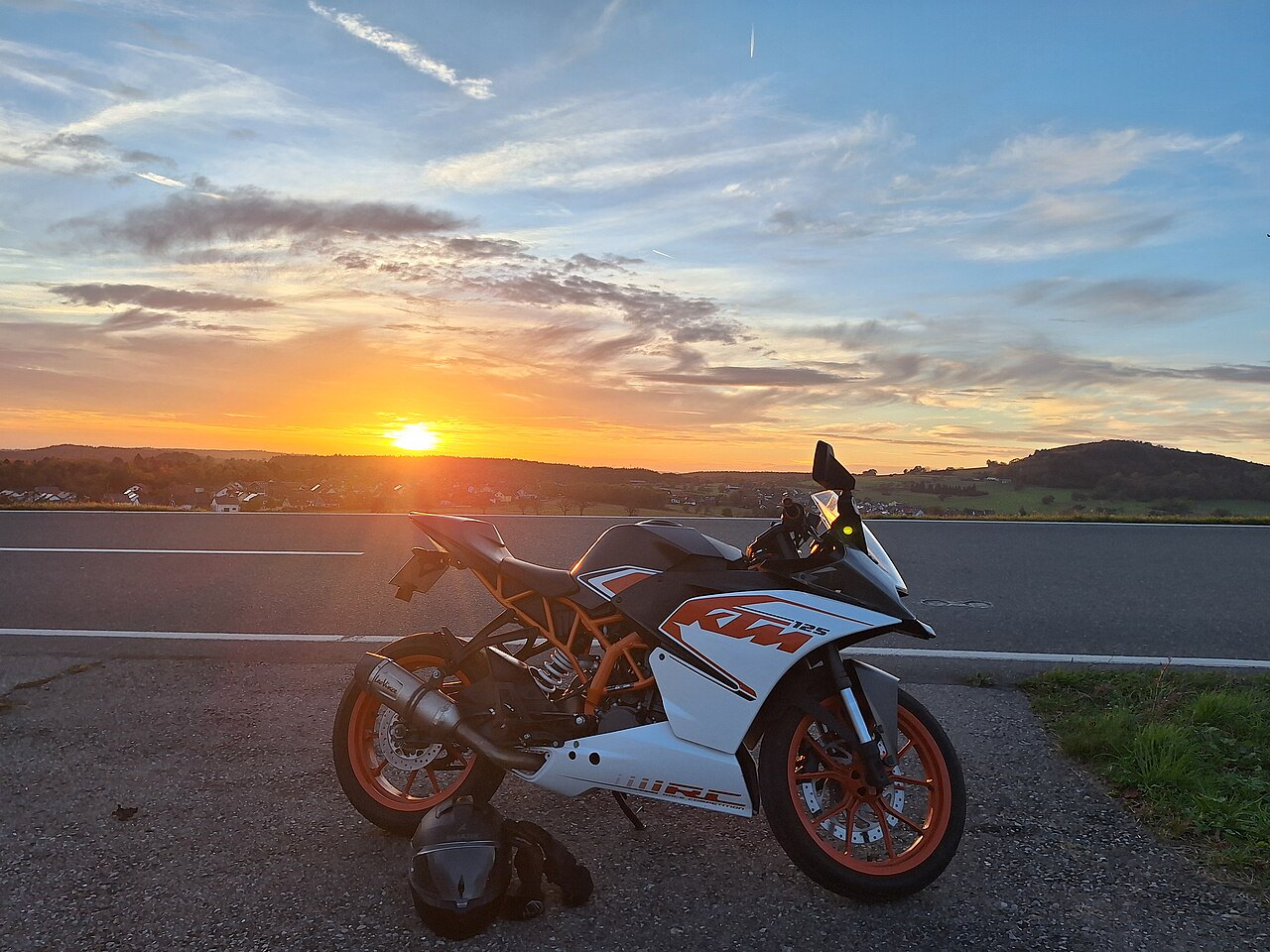 אופנוע KTM 125 RC לבן-כתום מונח על שפת כביש בזווית צד, ברקע שקיעה מרהיבה מעל שדה ורמות, עם קסדה ומעיל על הקרקע.