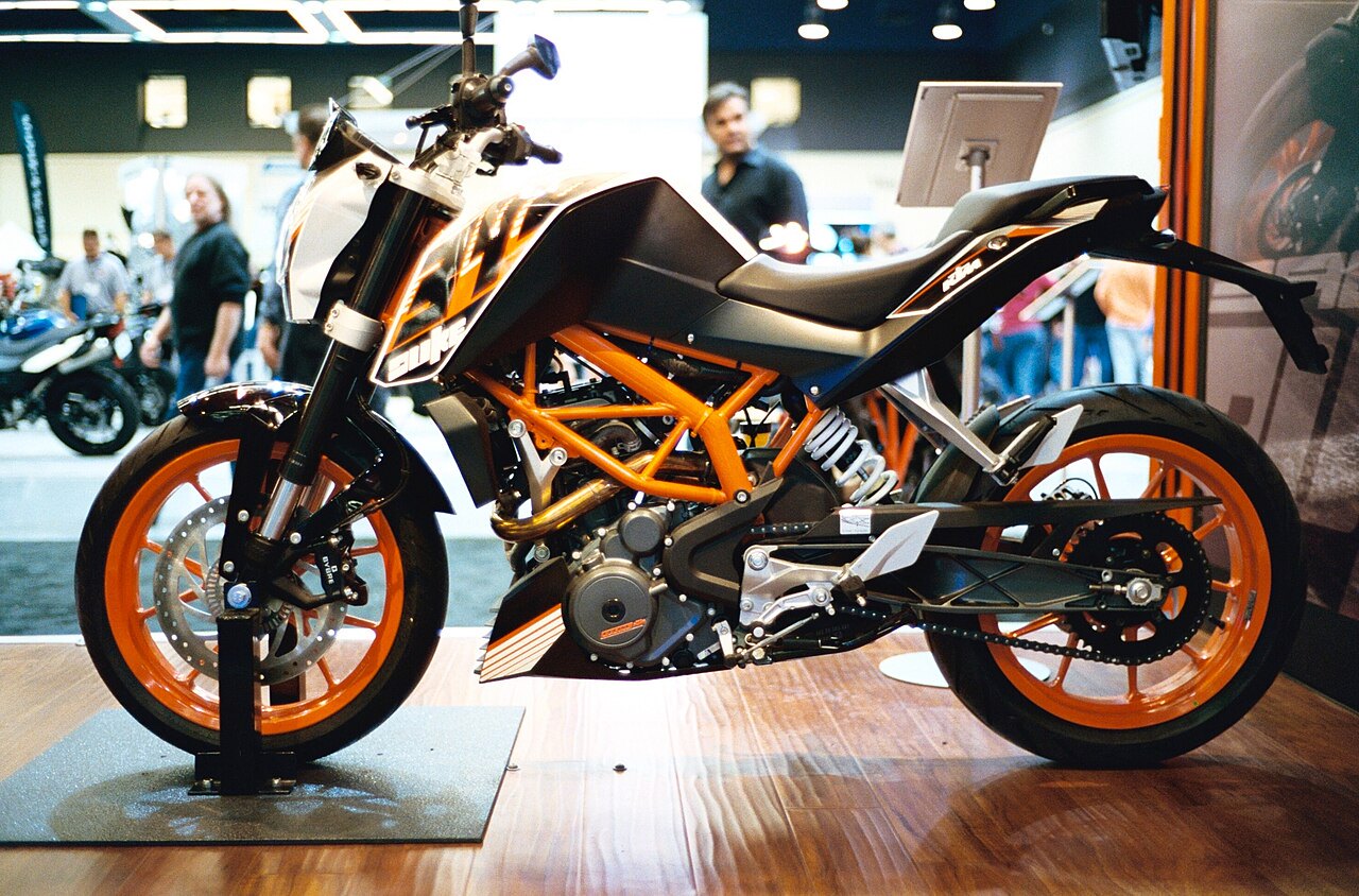 אופנוע KTM 1290 Super Duke R בצבע שחור עם מסגרת כתומה, צילום צד על מראה מוצב בפראט, רקע של תצוגה עם מבקרים